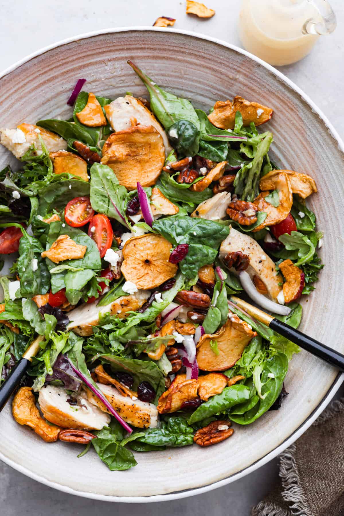 Top-down view of salad with chicken, greens, tomatoes, apples, nuts, and feta cheese in a stoneware bowl.