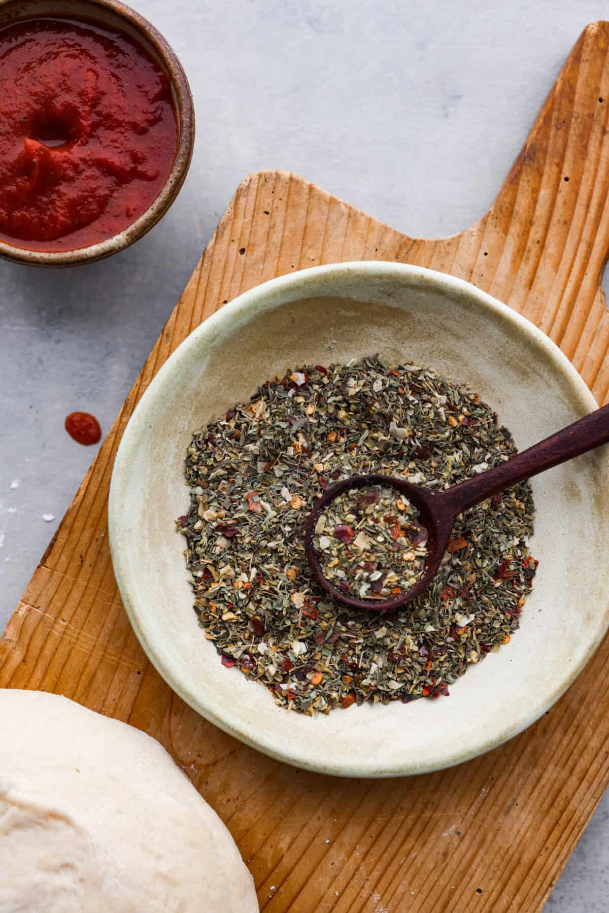 A small bowl of pizza seasoning on a wooden board.