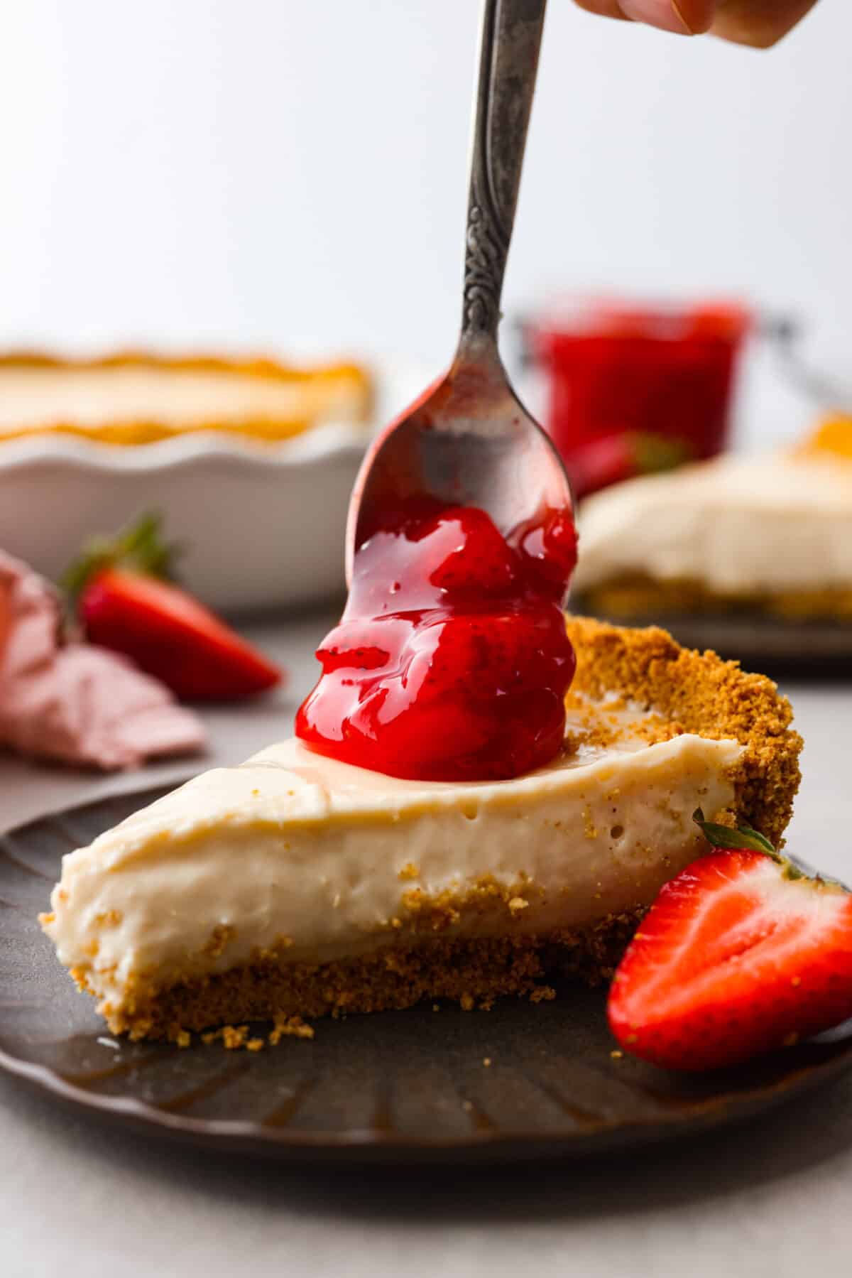 Strawberry pie filling being dolloped onto a piece of cheesecake. 