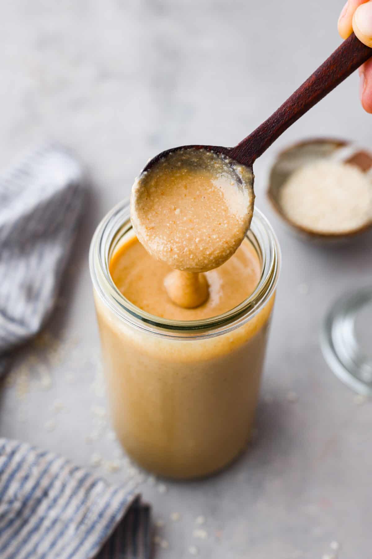 Top view of tahini in a glass jar with a wood spoon lifting up spoonful of tahini.