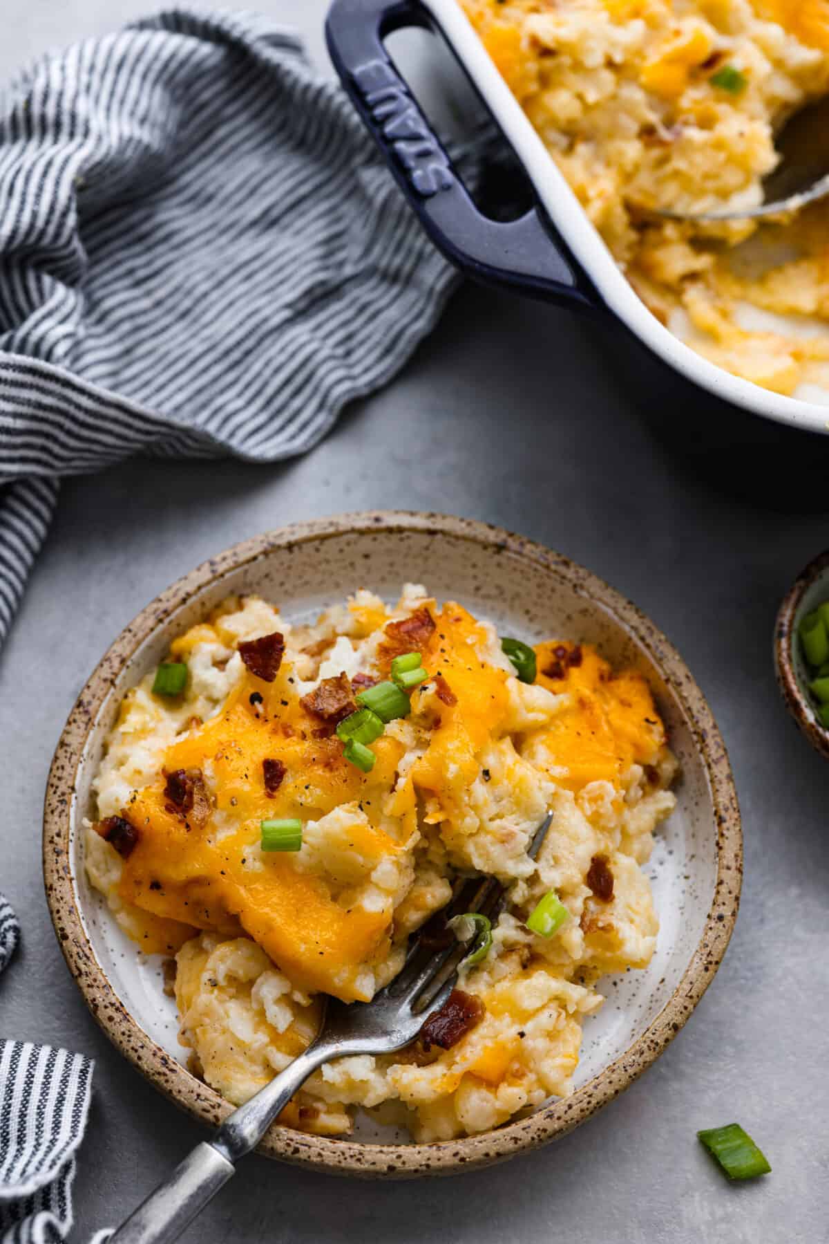 Top close view of a small plate of twice baked potato casserole. A fork is on the plate and the casserole dish next to it.