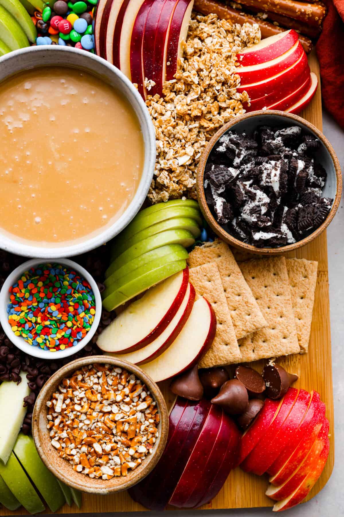 Closeup of caramel sauce in a white bowl as well as oreos, apple slices, m&ms, and granola.