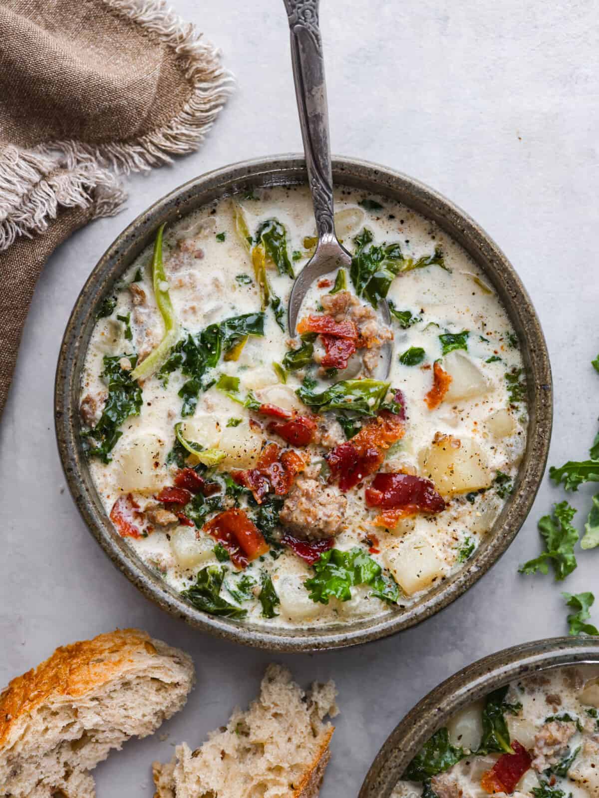 A bowl of soup with a silver spoon. There is some bread lying to the side of the bowl. 