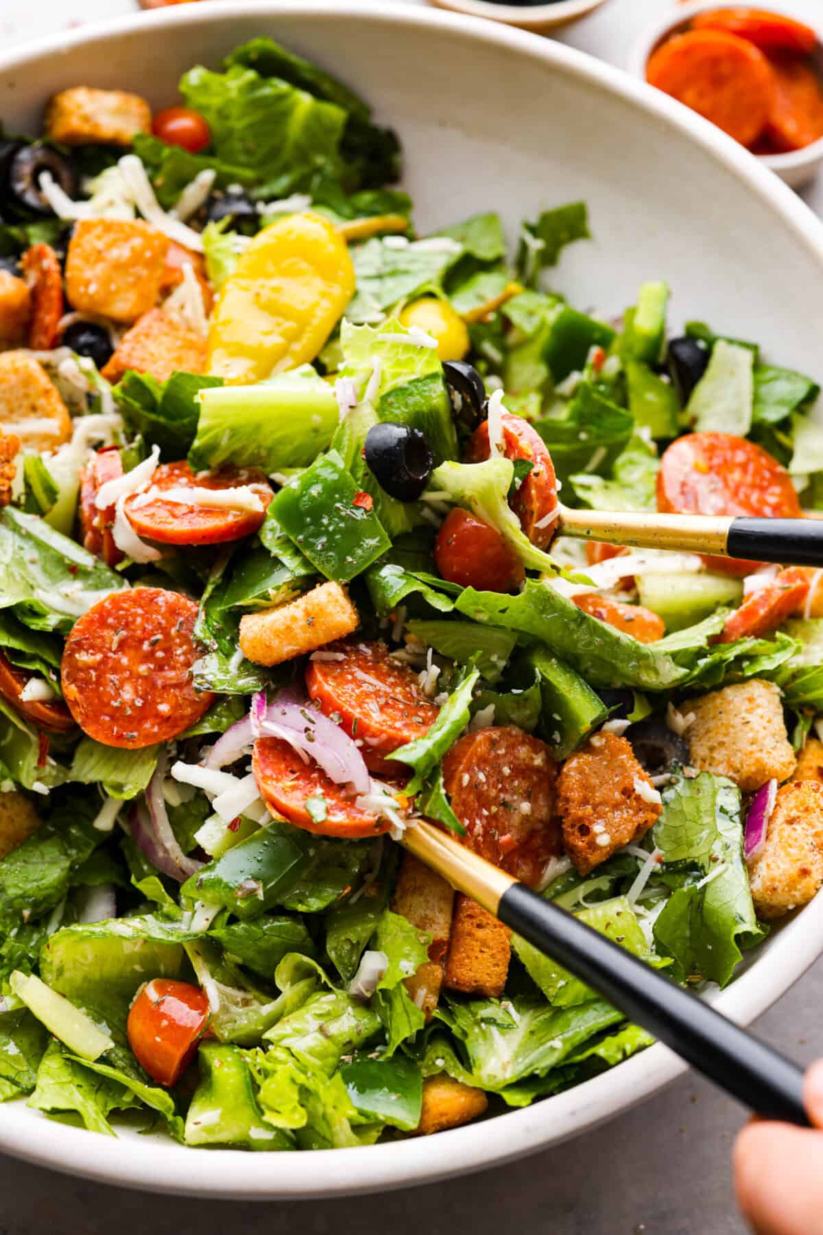 Pizza salad being tossed in a bowl with gold tongs. 