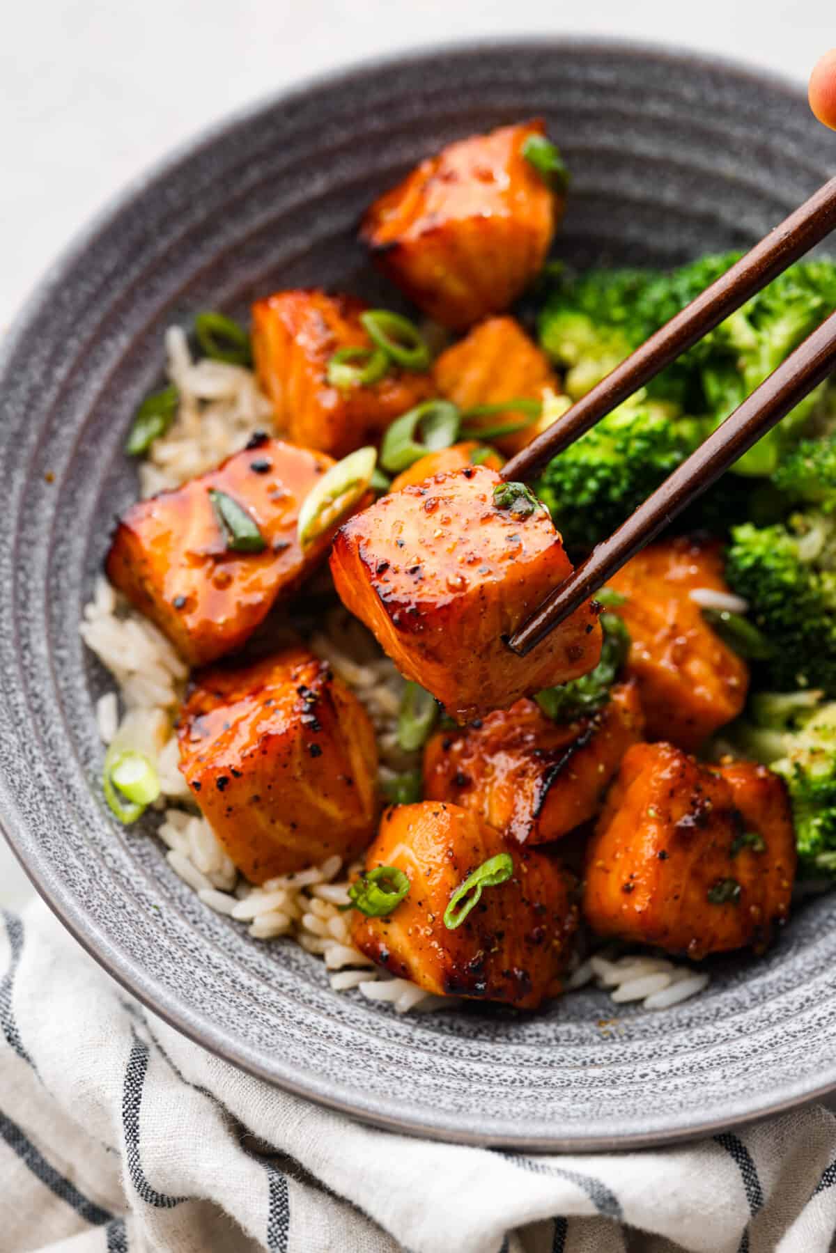 Close view of chopsticks lifting up a salmon bite in a bowl with rice and broccoli. 