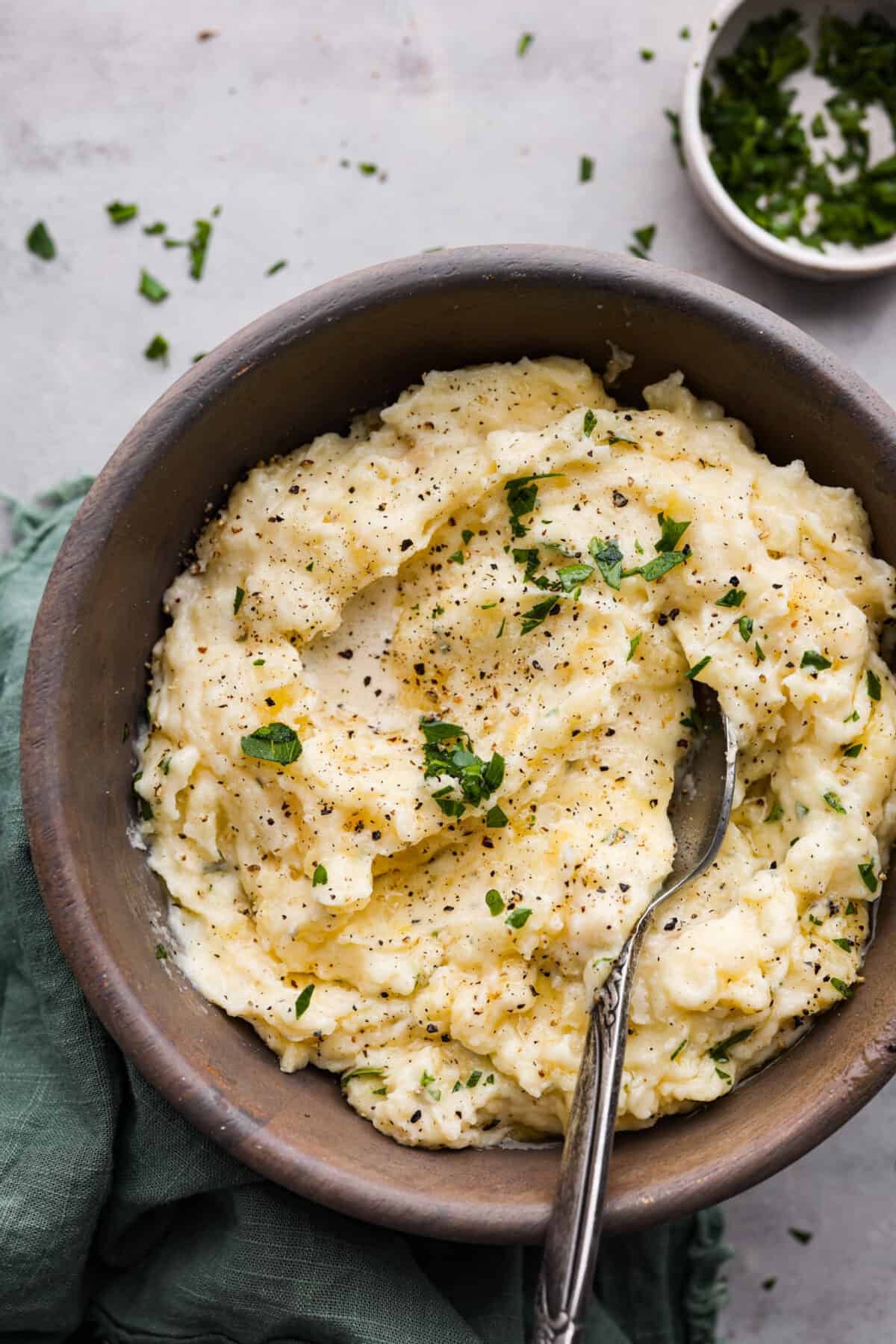 Top view of boursin mashed potatoes in a wood bowl and a silver soon. Garnished with fresh parsley, butter, and cracked black pepper.