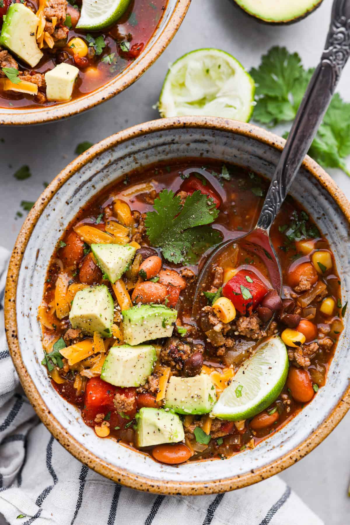 A bowl of taco soup, topped with chopped avocado, shredded cheese, cilantro, and a lime wedge.