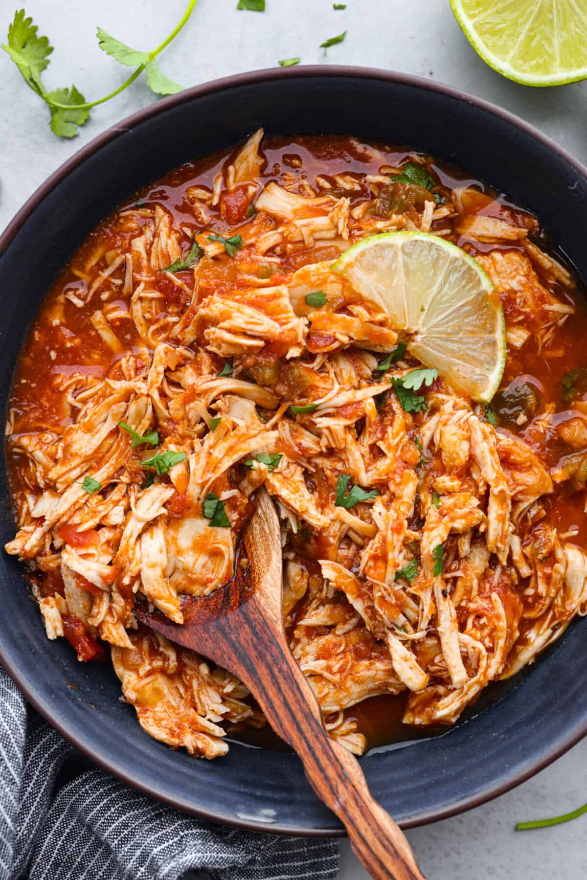 Chicken in a black bowl with a wood serving spoon and garnished with limes.