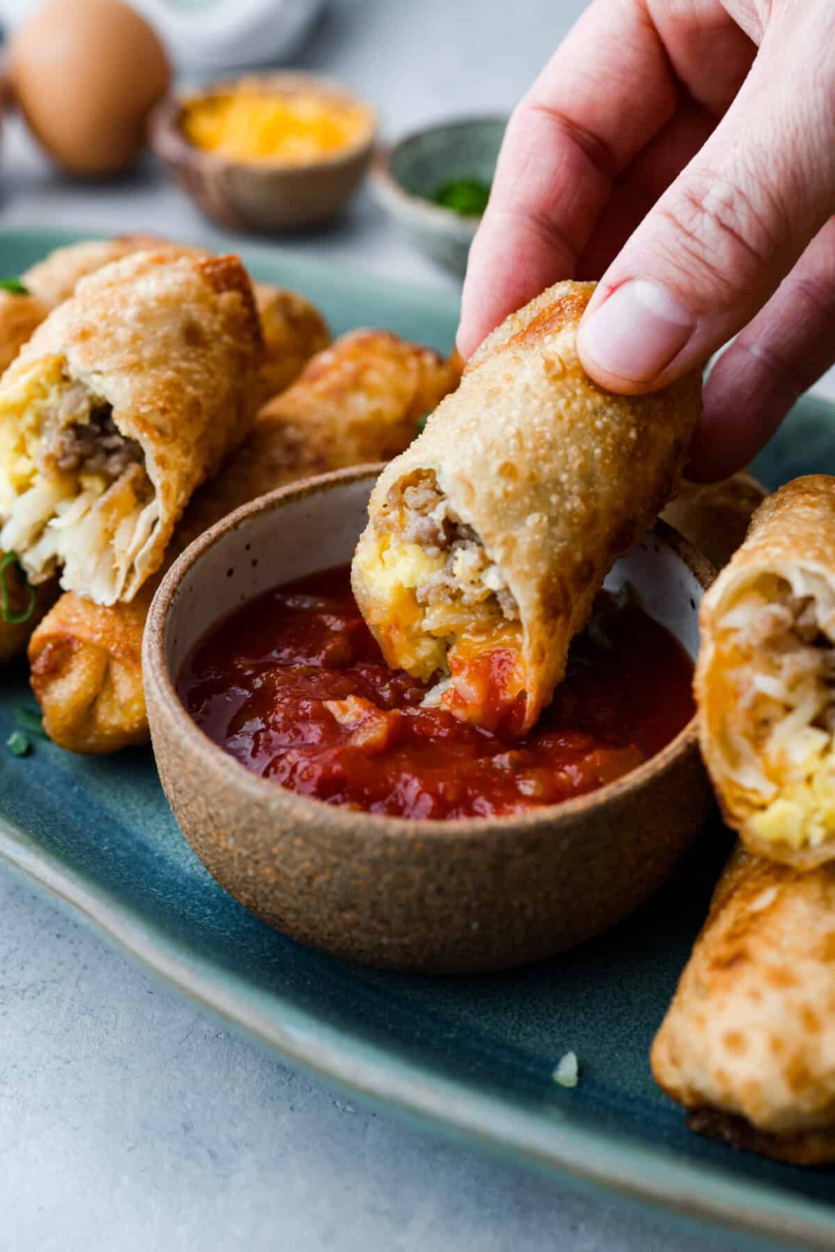 Side view of someone holding half of a breakfast egg roll dipping it into salsa. 