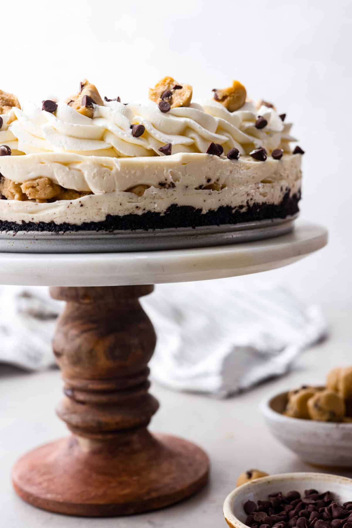 Side shot of completed no-bake cookie dough cheesecake on a cake stand. you can see all the layers and toppings. 