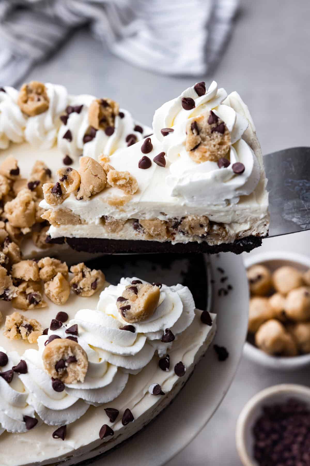 Slice of no-bake cookie dough cheesecake being taken out from the rest of the cheesecake. There is an Oreo crust, layers of creamy cheesecake filling and bits of cookie dough. Topped with more pieces of cookie dough mini chocolate chips and whipped cream.