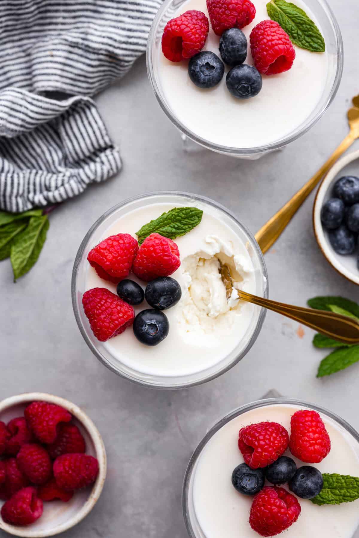 Overhead shot of a cup of Swedish cream with berries and mint on top, but a gold spoon sticking out of the top. There are other bowls filled with raspberries and blueberries nearby. 