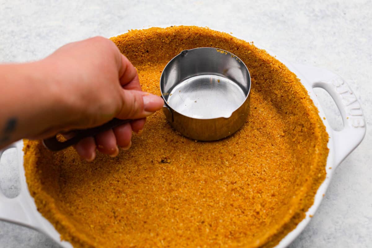 A crust being pressed into a pie pan with a measuring cup. 