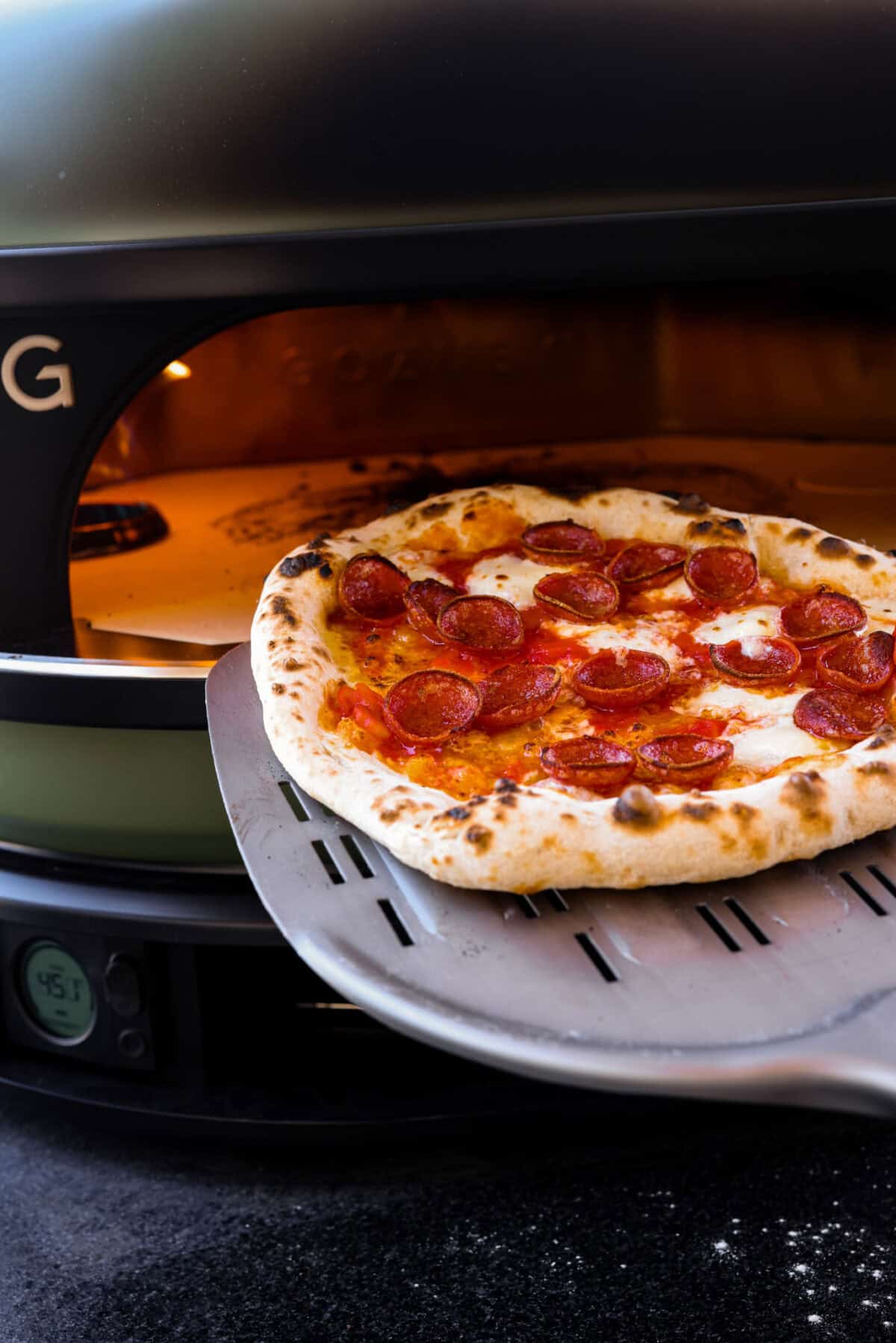 Close view of a pizza spatula removing cooked wood-fired pizza from the oven.