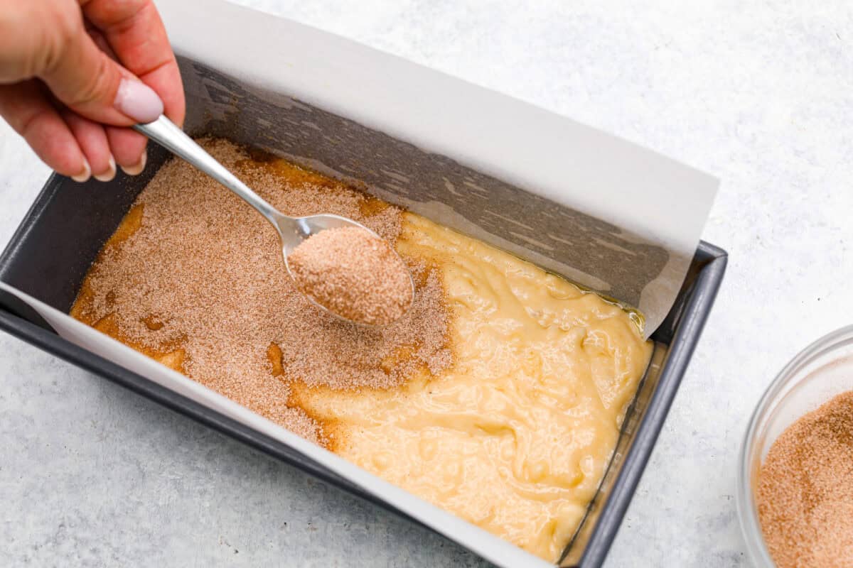 Overhead shot of banana bread batter in pan while someone sprinkles cinnamon and sugar mixture over the top. 