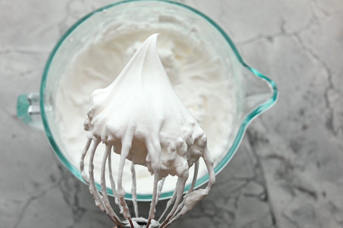 Overhead shot with bowl of whipped egg whites in the background with a stand mixer whisk attachment in the foreground with peaked egg whites. 