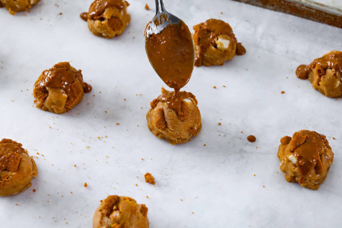 Angle shot of a spoon drizzling melted cookie butter over cookie dough balls on cookie sheet. 