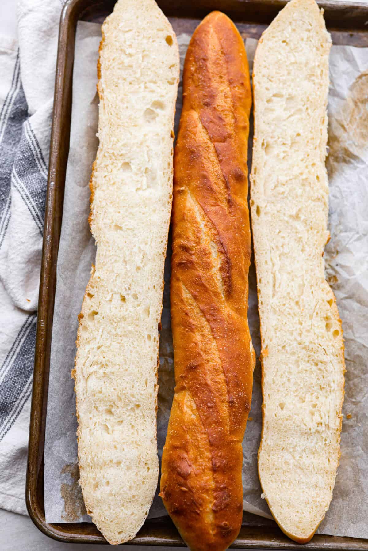 Overhead shot of baked French baguette loaves on a baking sheet, one is cut in half the long way. 