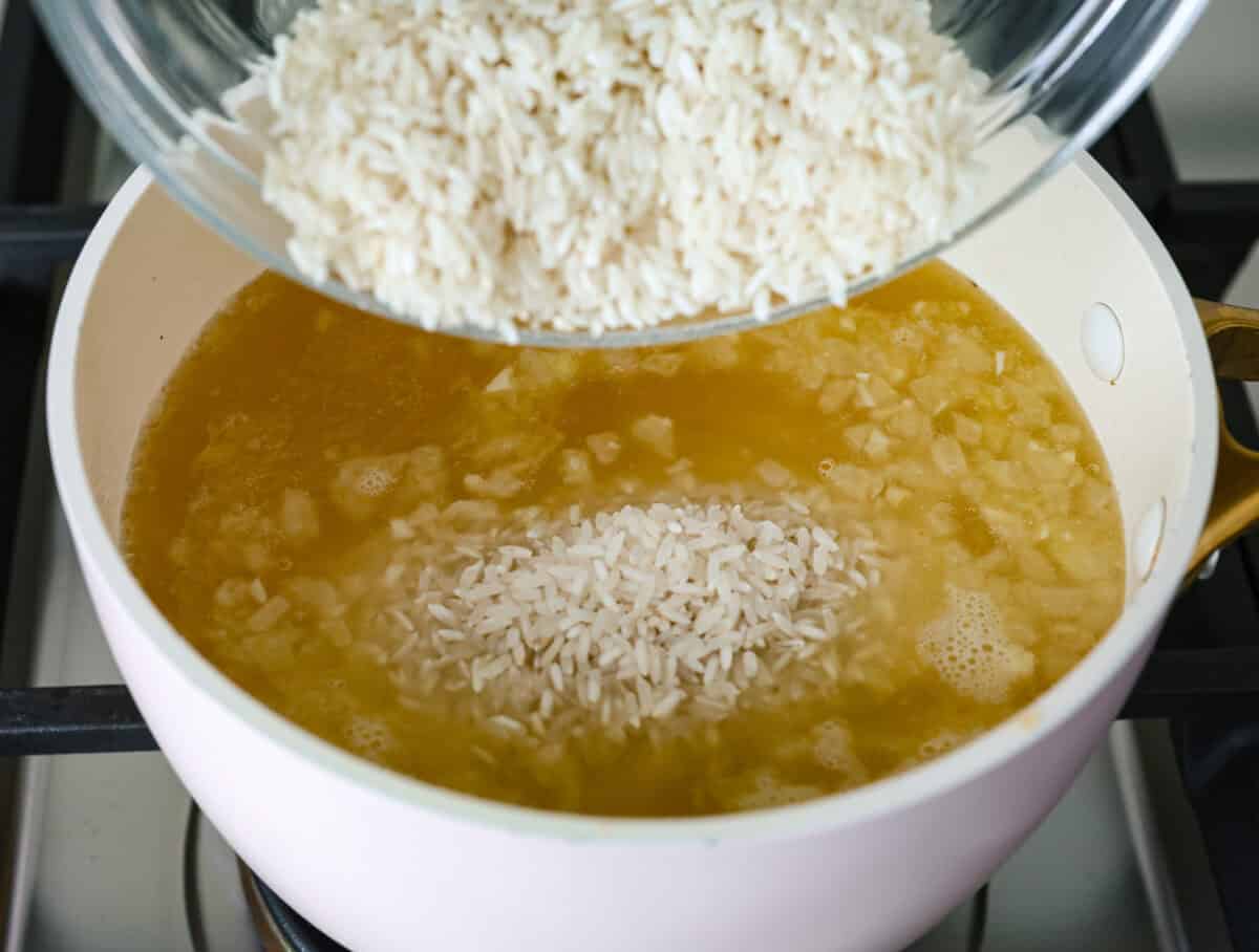 Overhead shot of someone pouring rice into the broth, butter, onion and garlic mixture in the pot. 