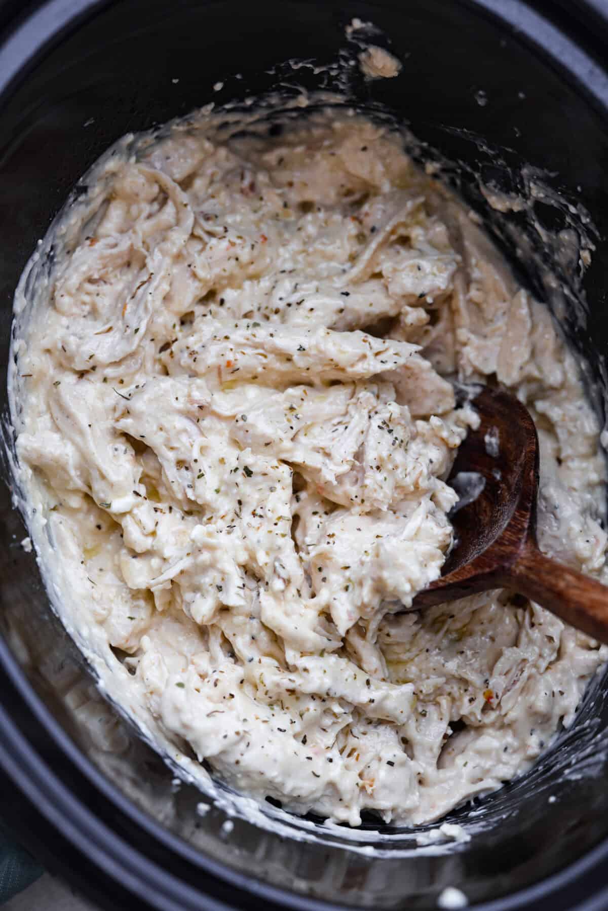 Overhead shot of cooked Italian cream cheese chicken in crockpot with wooden spoon. 