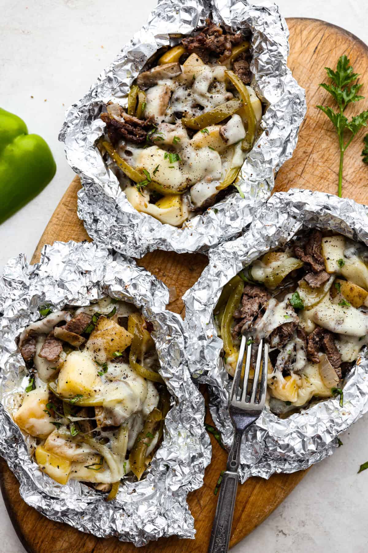 Overhead shot of 3 individual Philly cheesesteak foil packs open on a wood cutting board. 