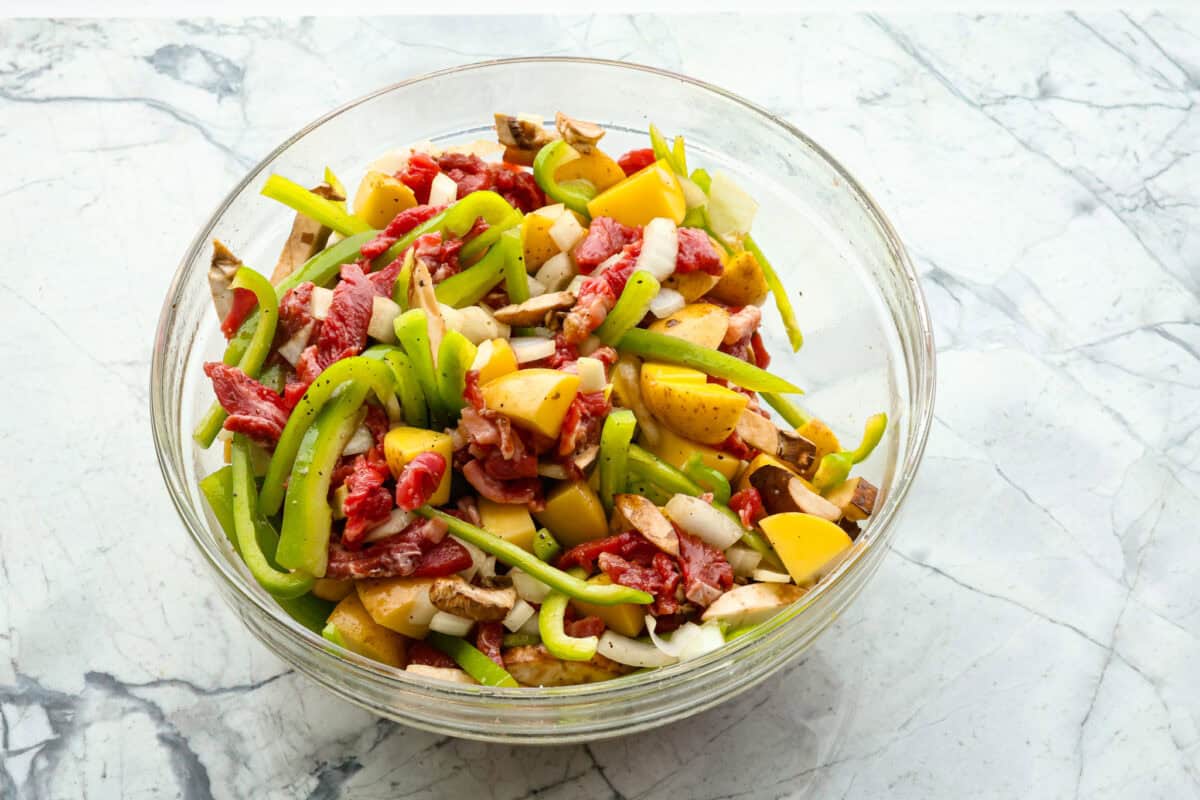 overhead shot of raw steak, peppers, onions, seasonings and potatoes in glass bowl. 