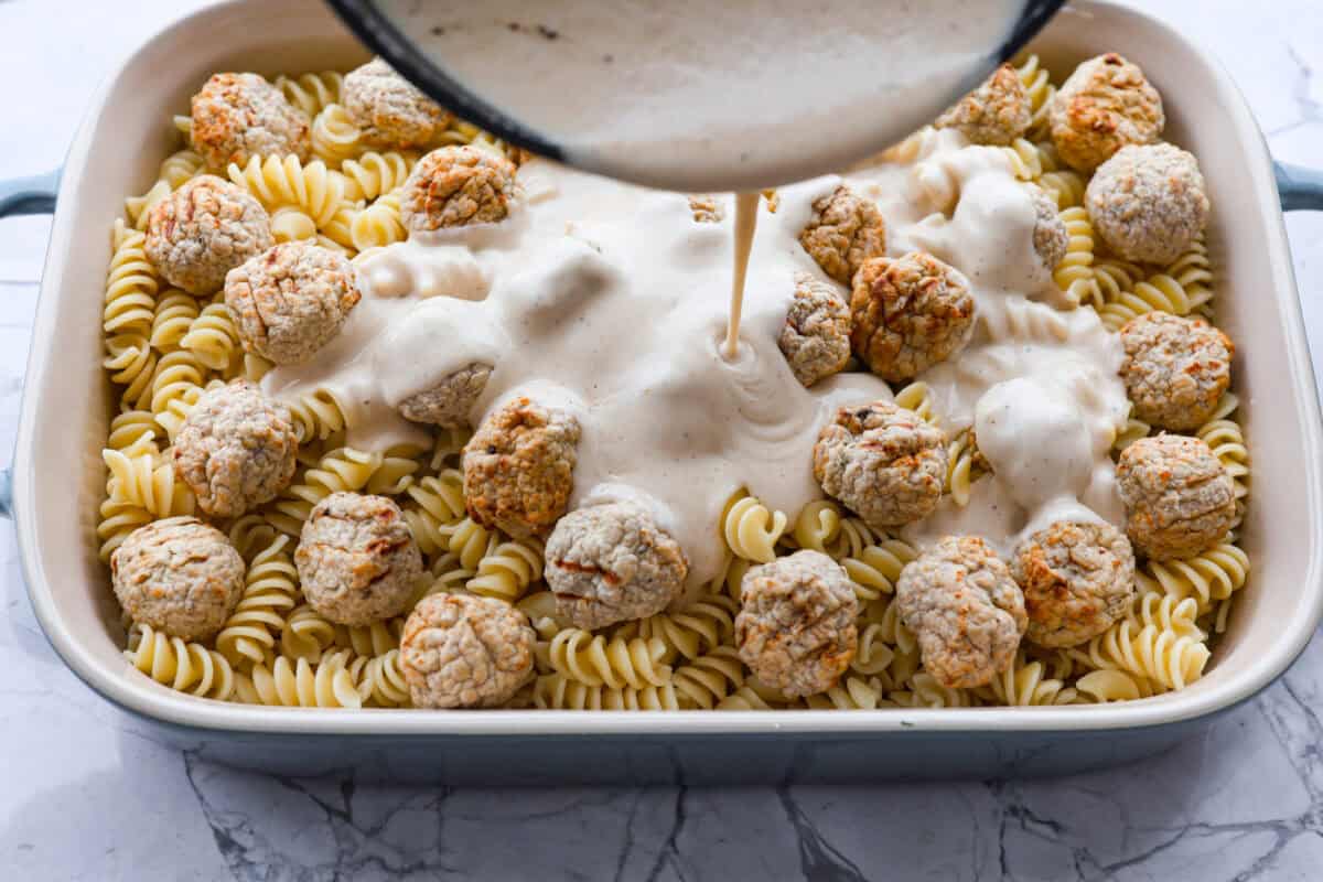 Overhead shot of someone pouring sauce over pasta and meatballs in a casserole dish. 