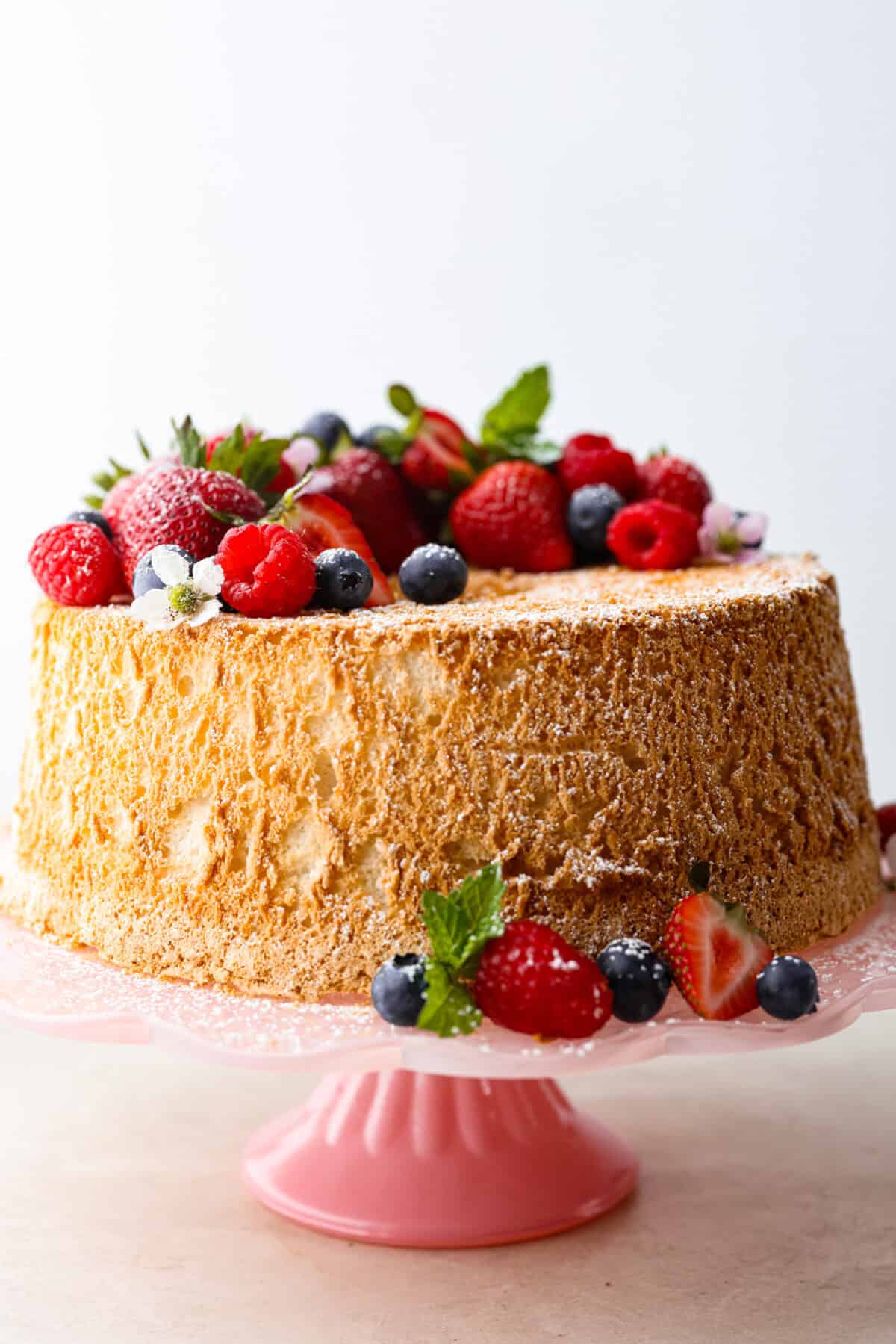 Side shot of whole angel food cake with berries and blossoms dusted with powdered sugar. 