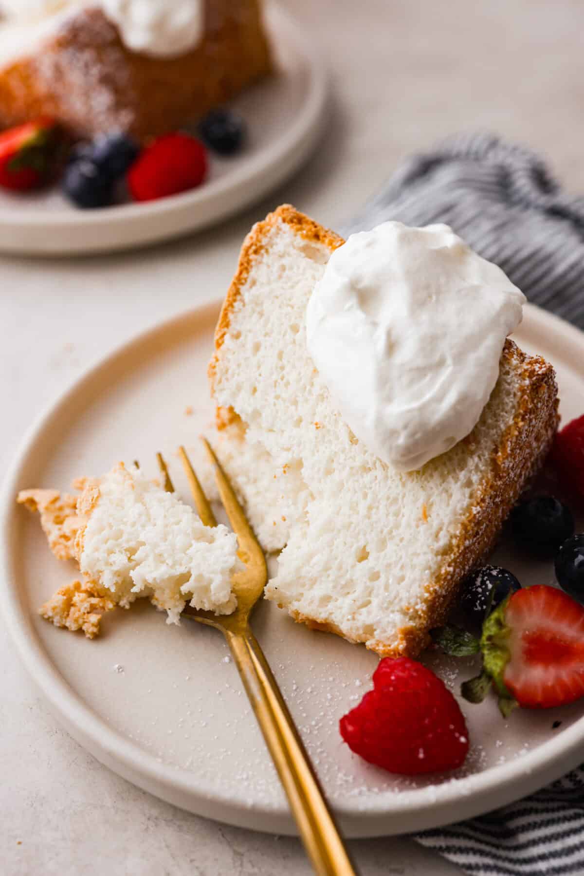 Angle shot of plated angel food cake with dollop of whipped cream on top and berries on the side, next to a gold fork.