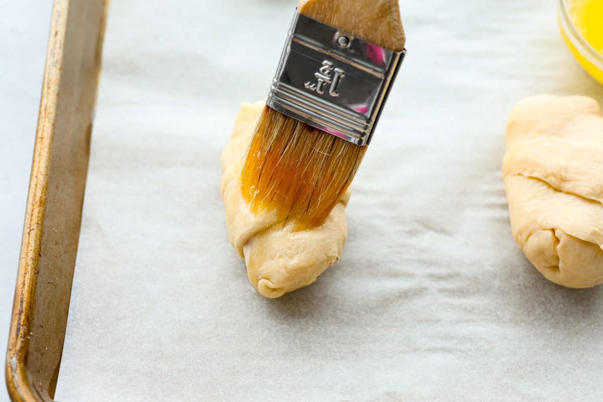 Angle shot of butter being spread on top of crescent roll. 