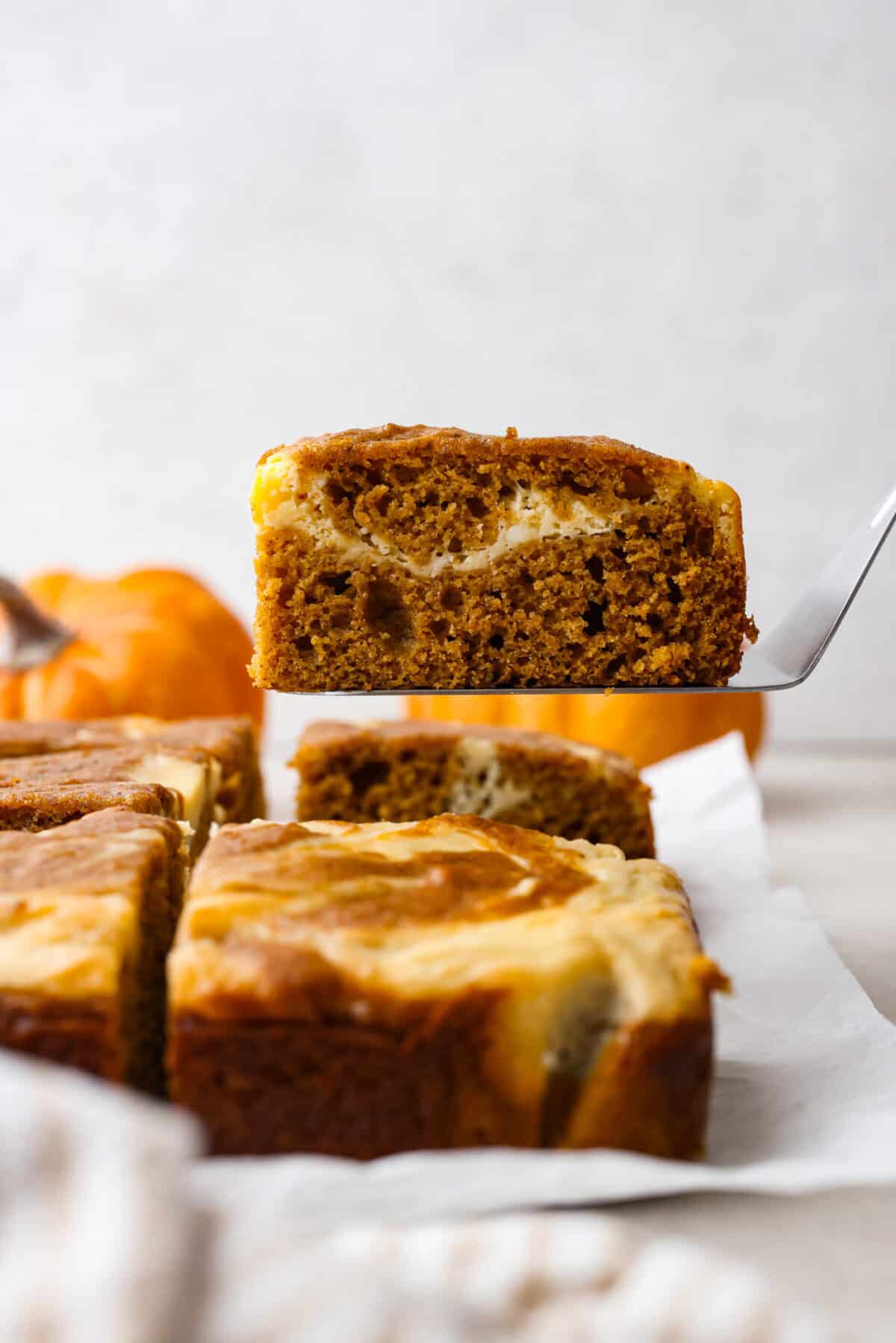 Side shot of a pumpkin roll bar on a spatula being lifted up off a counter, that has parchment paper with other bars on it and small pumpkins in the background. 