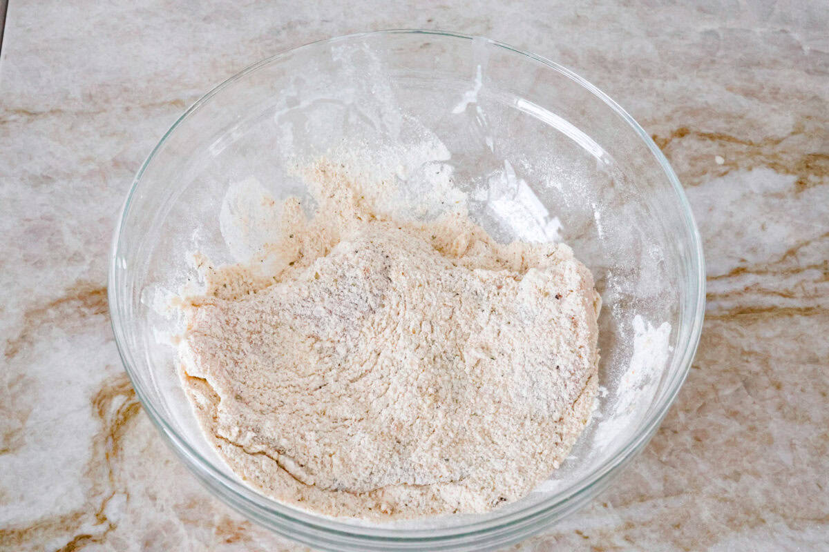 Overhead shot of chicken cutlet being dredged in breading mixture. 