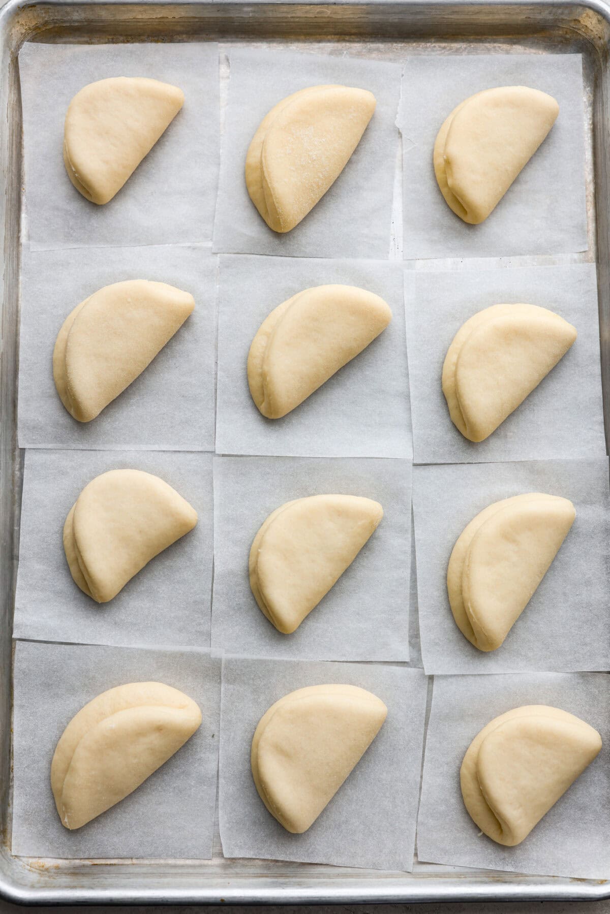 Overhead shot of bao bun dough circles folded in half on individual parchment papers on a cookie sheet. 