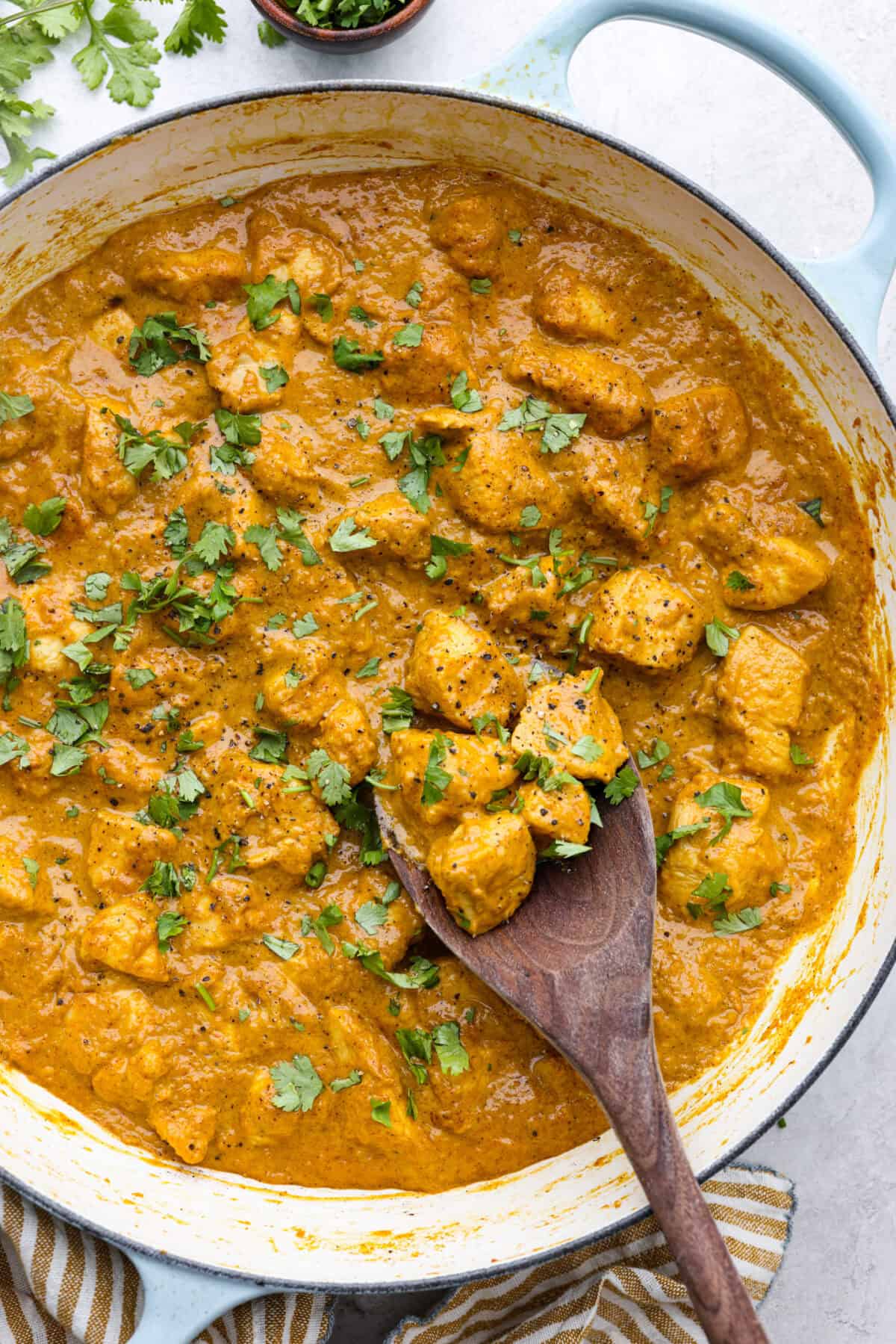 Overhead shot of chicken korma in a skillet with wooden spoon and garnished with cilantro. 