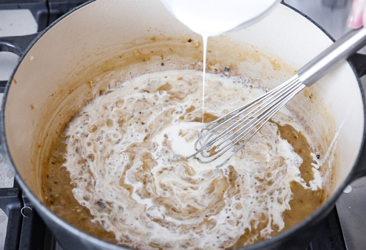 Angle shot of someone slowly pouring in the chicken broth and heavy cream into the roux in a pot. 