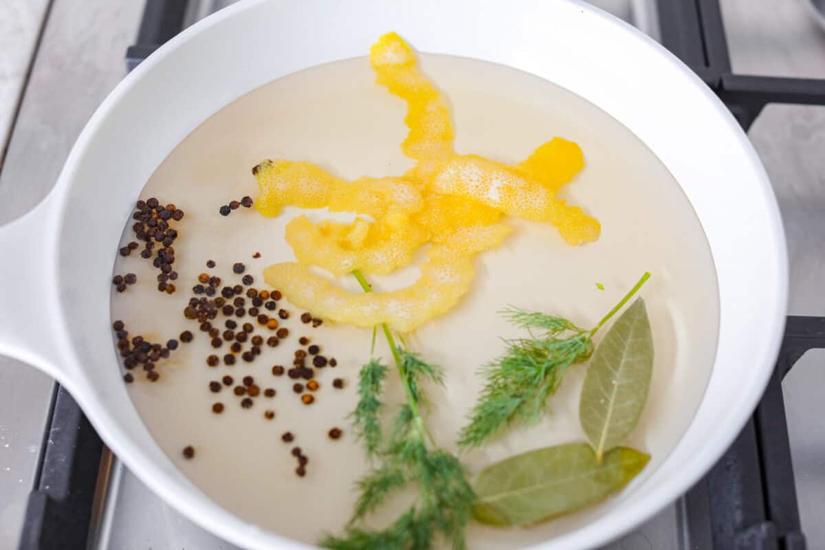 Overhead shot of white wine with lemon peal, fresh herbs and peppercorns in a skillet on the stove. 