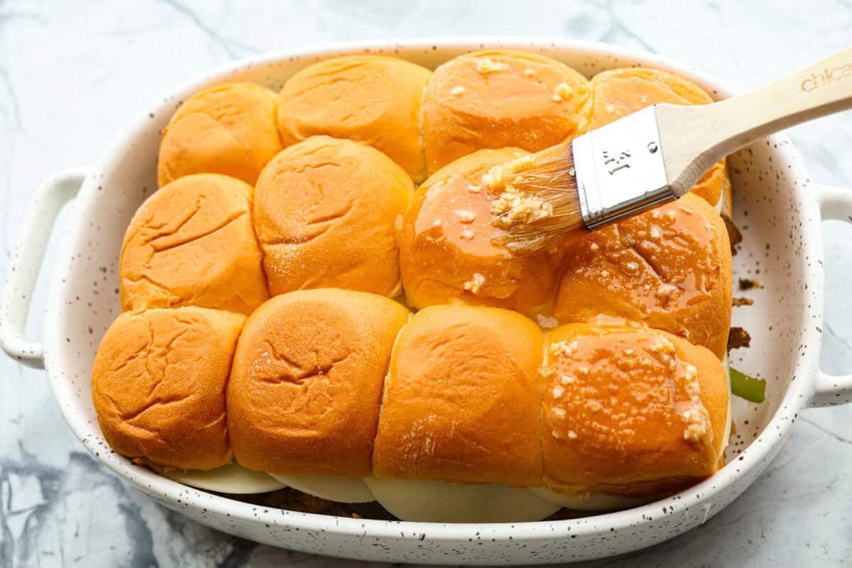 Overhead shot of someone basting the top of the Philly cheesesteak slider buns with butter and seasoning mixture. 