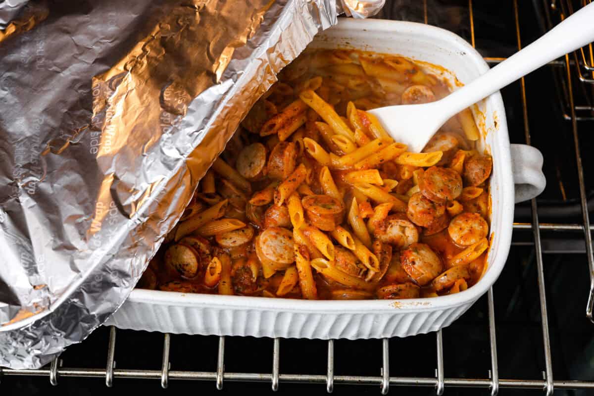 Angle shot of someone lifting the tin foil off the pan and stirring the noodle bake. 