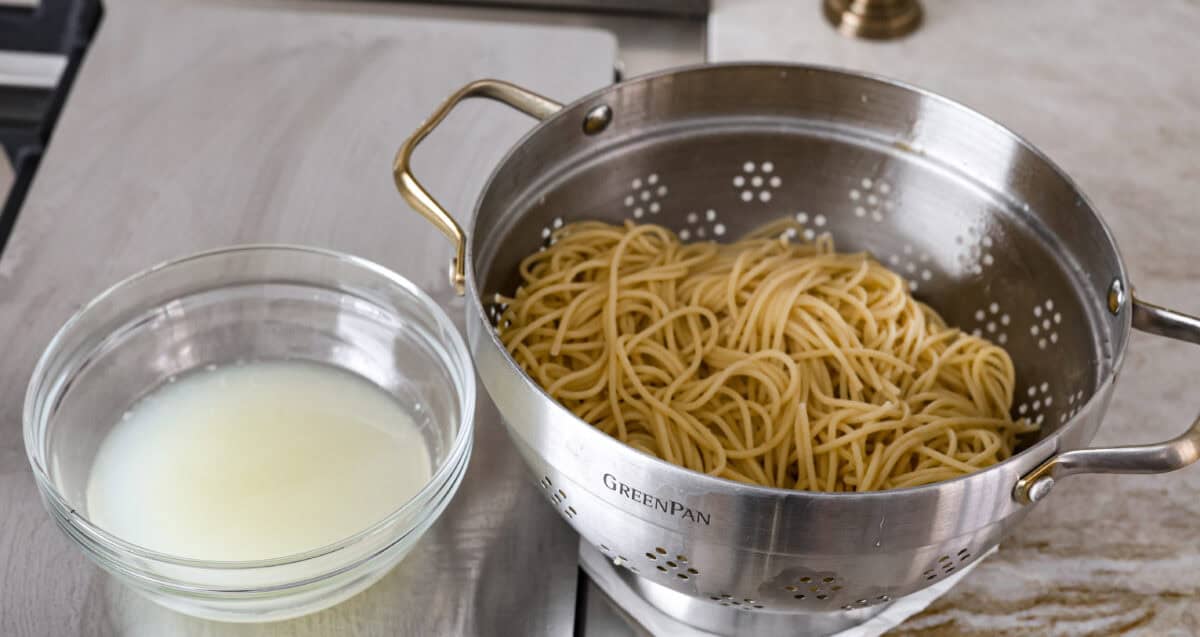 Angle shot of cooked spaghetti noodles in a colander with pasta water next to it in a separate bowl. 