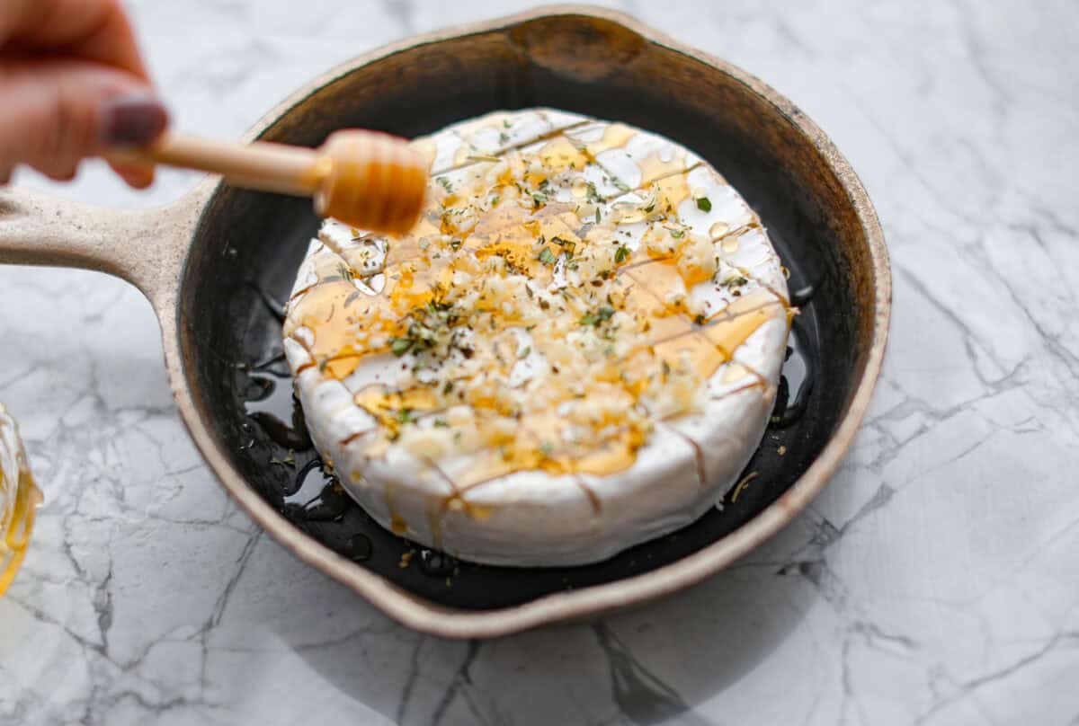 Angle shot of someone drizzling honey over garlic and herbs on the circle of cheese in a skillet. 