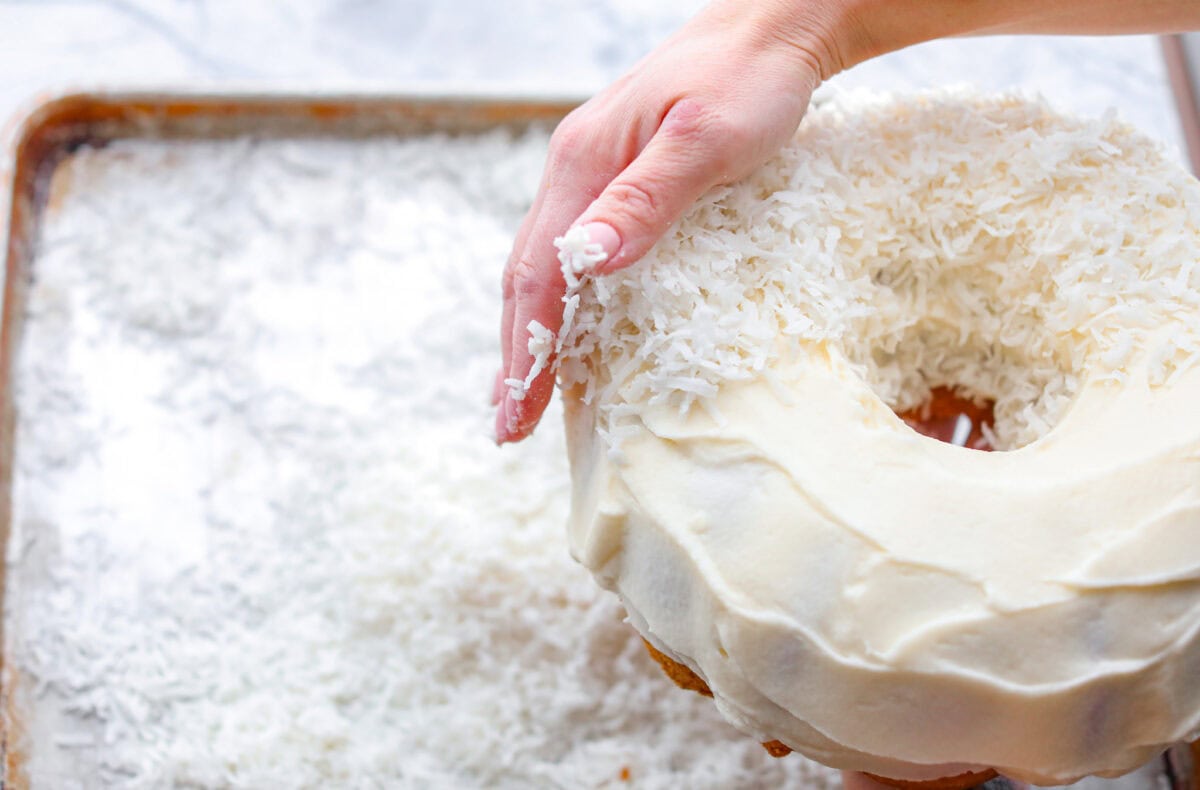 Angle shot of someone pressing shredded coconut flakes onto the frosted cake. 