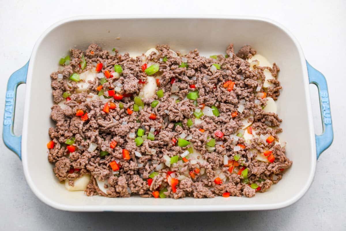 Overhead shot of beef and veggie mixture spread over the potatoes in the bottom of a baking dish. 