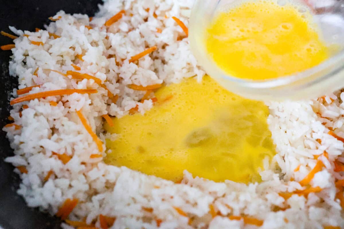 Close up shot of someone pouring whisked eggs into a space made in the middle of the rice and veggies in the skillet. 