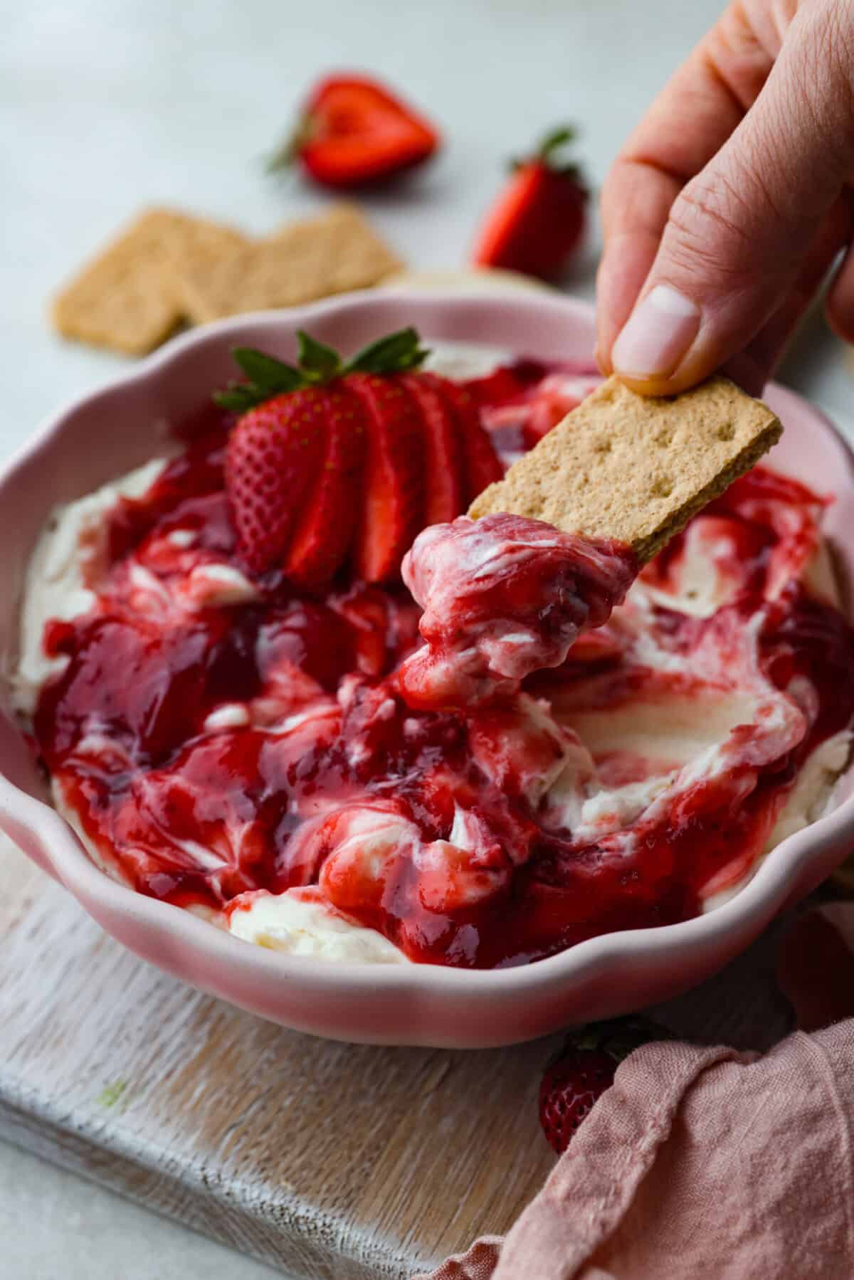 Angle shot of someone dipping a graham cracker into the strawberry cheesecake dip. 