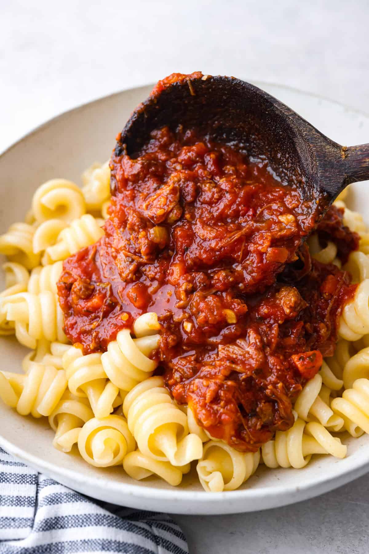 Side shot of someone pouring a scoop of slow cooker Sunday sauce over a plate of cooked pasta. 