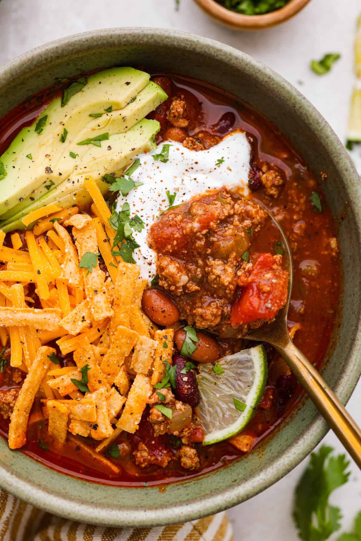 Close up shot of a bowl of turkey chili with avocado, lime, cheese sour cream and tortilla strips for garnish. 